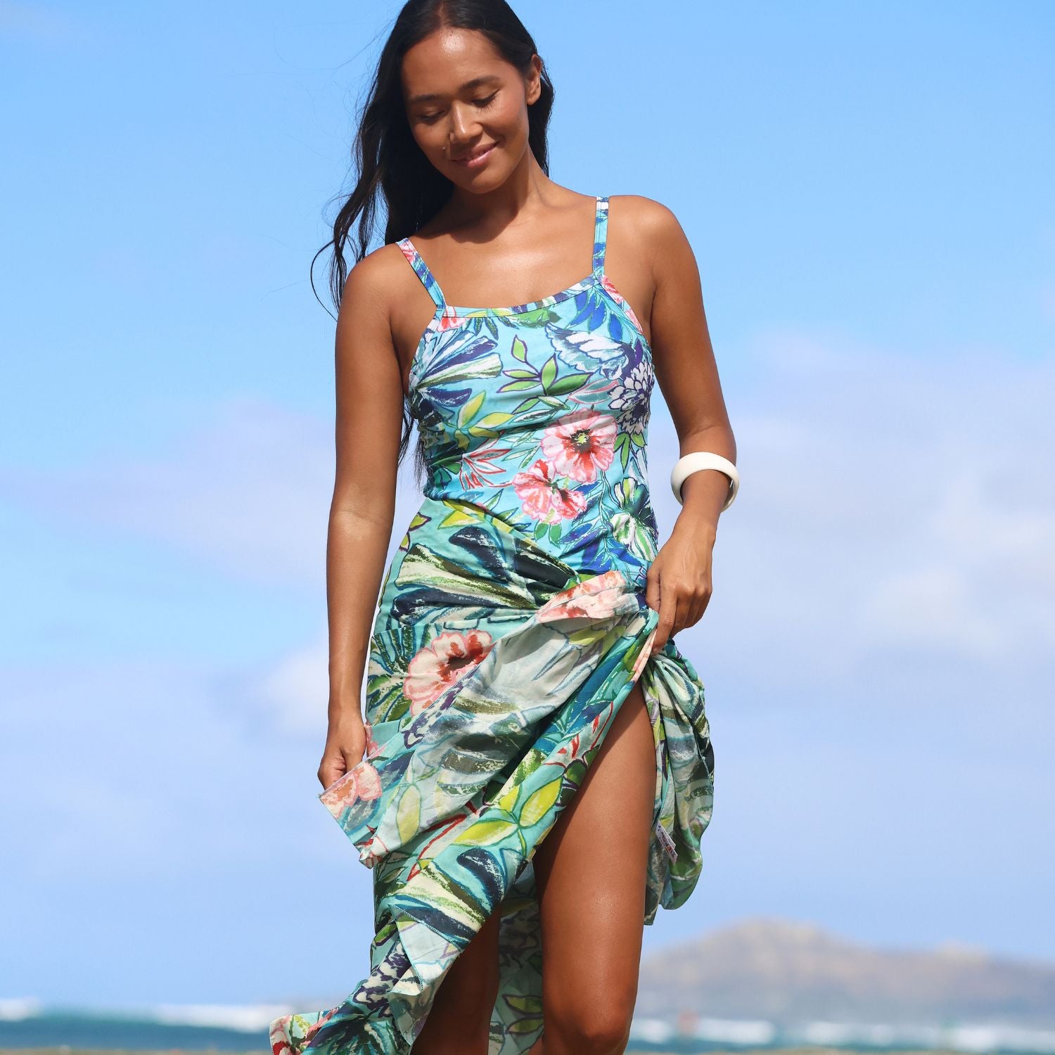 A woman wearing a tropical floral print high-slit dress, standing on a beach with a blue sky and distant mountains in the background.