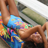 A woman reclining on a colorful tropical print beach towel or blanket next to a white wooden bench, wearing a bright blue one-piece swimsuit with a multicolor floral and leaf pattern.