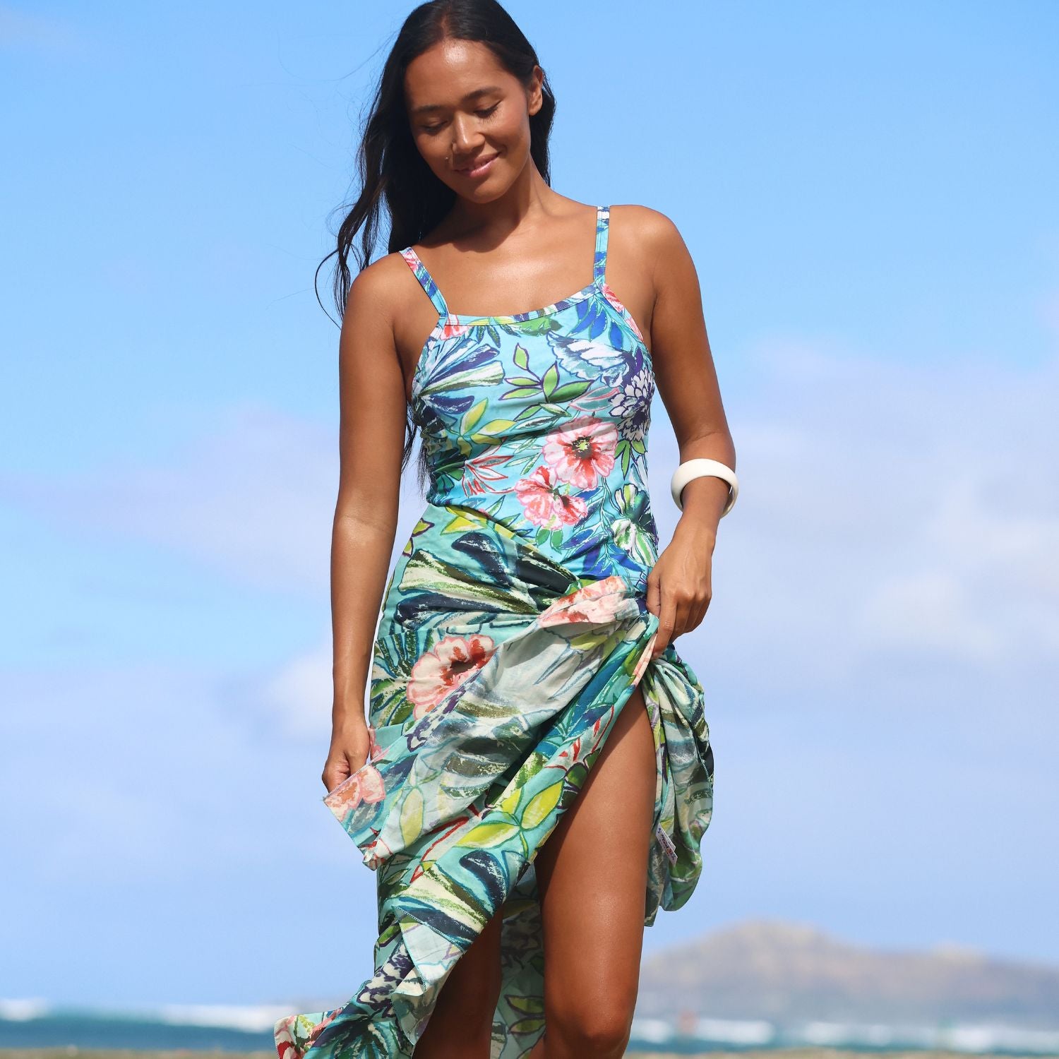 A woman wearing a tropical floral print high-slit dress stands on a beach with the ocean and distant mountains in the background. The dress features a vibrant mix of blue, green, pink, and yellow tones in a leaf and hibiscus flower pattern.