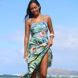 A woman wearing a tropical floral print high-slit dress stands on a beach with the ocean and distant mountains in the background. The dress features a vibrant mix of blue, green, pink, and yellow tones in a leaf and hibiscus flower pattern.