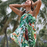 A woman wearing a tropical print halter wrap dress with green palm leaves and orange flowers, posing outdoors with palm trees in the background.