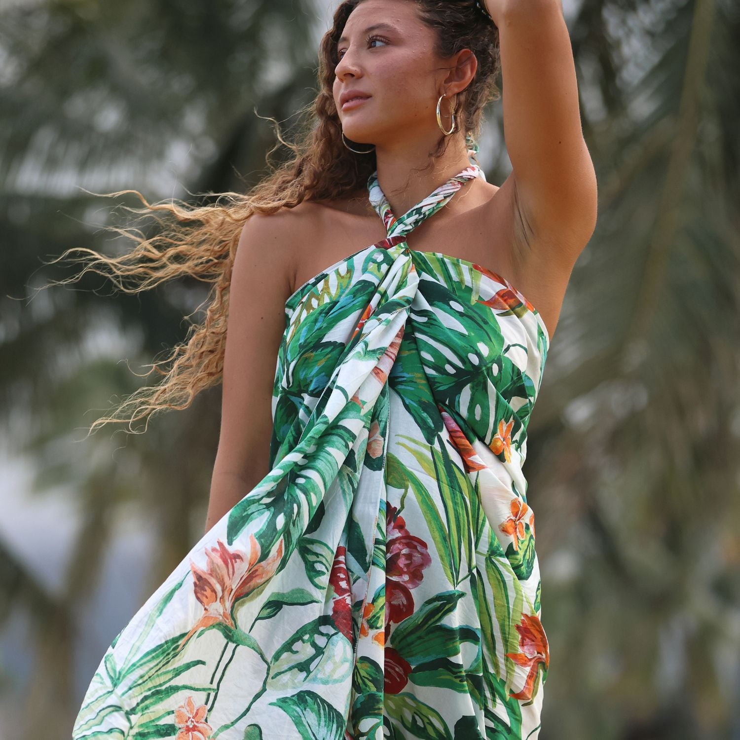 A woman wearing a halter-neck tropical print dress with green monstera leaves and orange flowers on a white background, photographed outdoors with blurred palm trees.