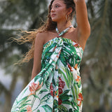 A woman wearing a halter-neck tropical print dress with green monstera leaves and orange flowers on a white background, photographed outdoors with blurred palm trees.