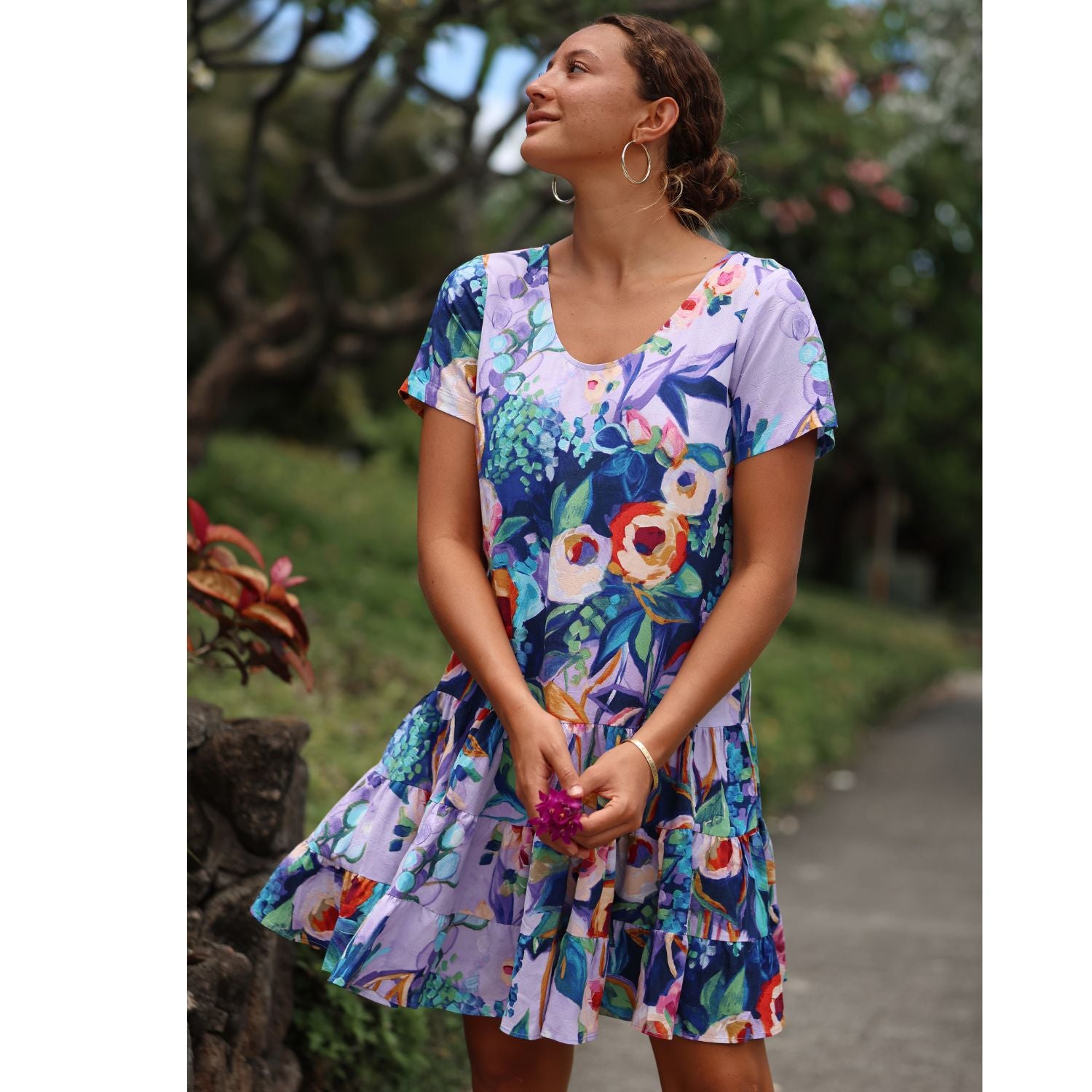 A woman wearing a short-sleeve tiered skater dress with a colorful floral print in shades of purple, blue, green, and orange, standing outdoors on a path with trees in the background.