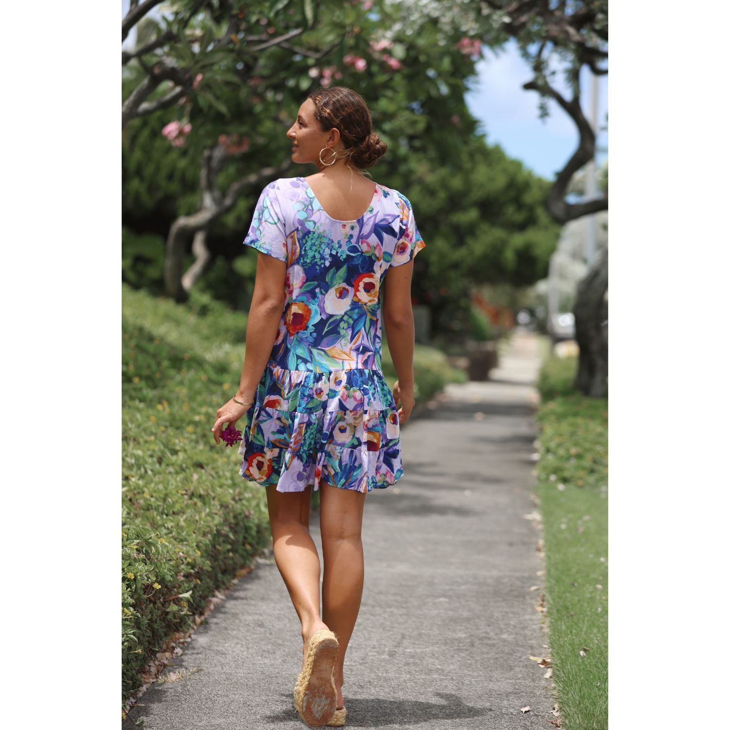 A woman wearing a colorful floral print short-sleeve dress walks along a paved garden path, photographed from behind.