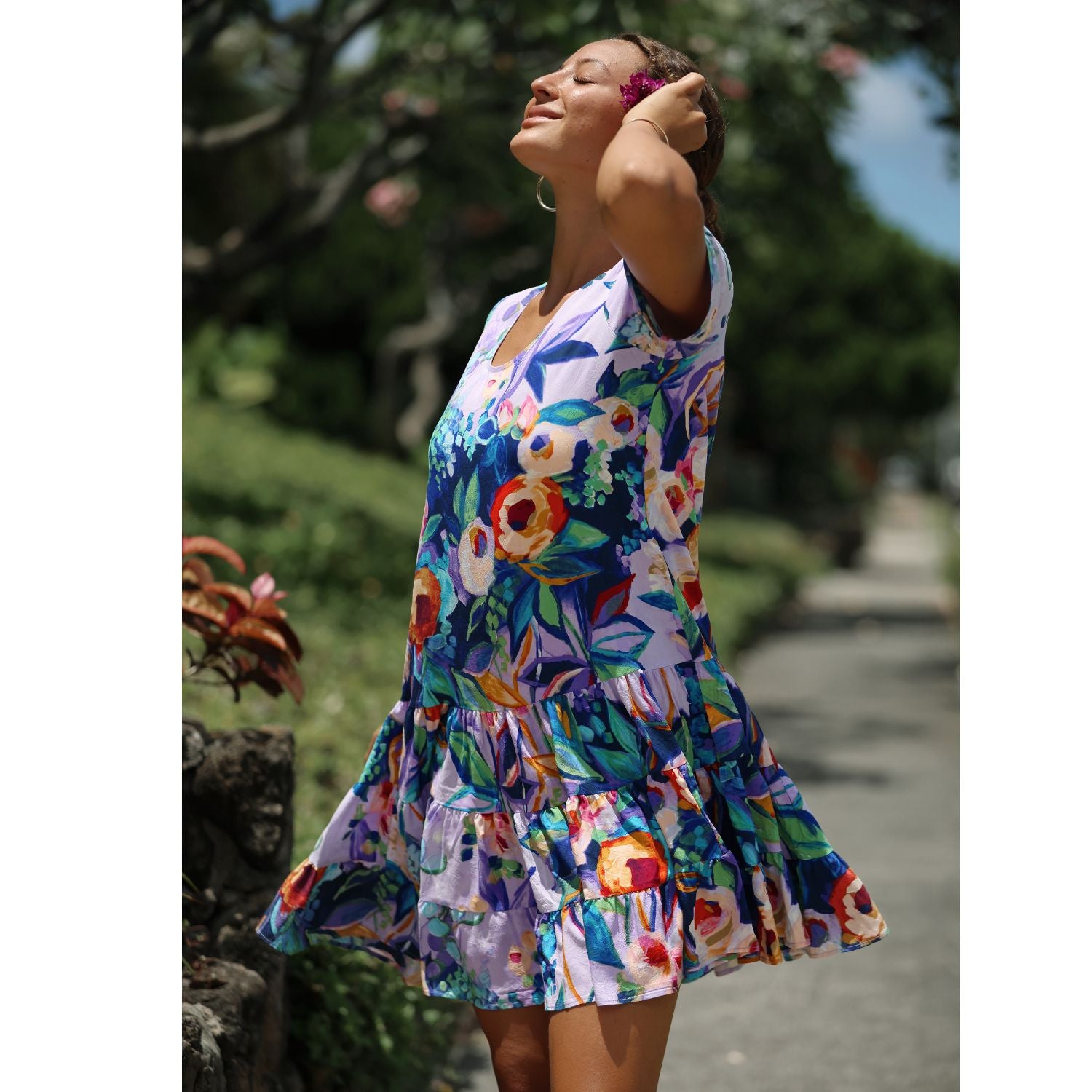 A woman wearing a colorful floral tiered dress stands outdoors on a path with trees and greenery in the background.