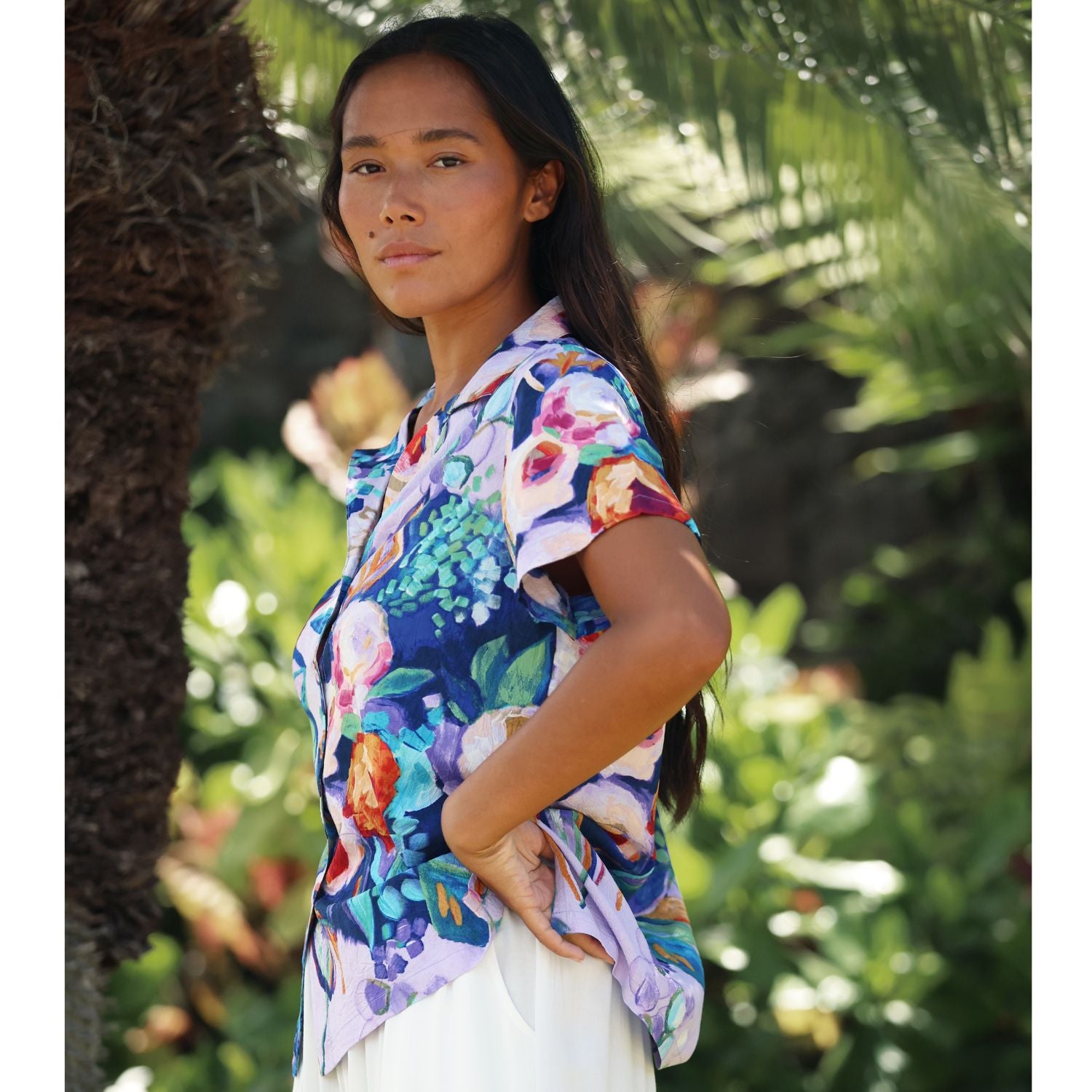 A woman wearing a short-sleeve button-up shirt with a vibrant multicolor floral print, posed outdoors against a blurred background of green foliage and palm trees.