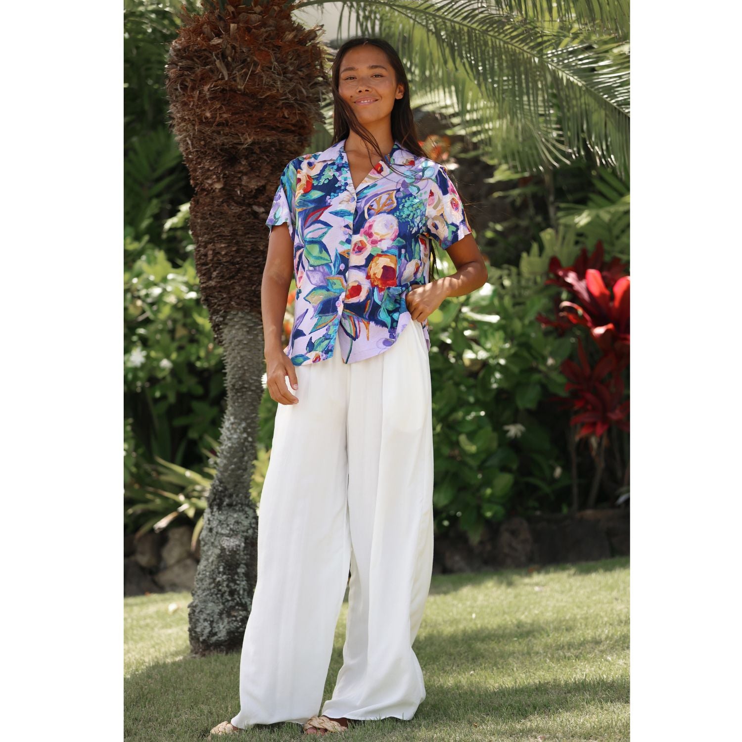 A woman stands outdoors in a tropical garden setting, wearing a short-sleeve button-up shirt with a colorful floral print and white wide-leg pants.