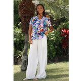 A woman stands outdoors in a tropical garden setting, wearing a short-sleeve button-up shirt with a colorful floral print and white wide-leg pants.