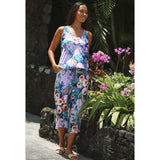 A woman wearing a sleeveless floral maxi dress with a matching headband, standing outdoors next to a stone wall and tropical plants with purple flowers.