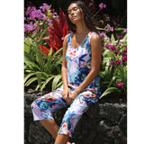 A woman wearing a sleeveless, knee-length jumpsuit with a colorful abstract floral print sits on a dark stone wall surrounded by lush green plants and purple flowers.
