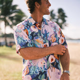 A man wearing a short-sleeve button-up Hawaiian shirt with a colorful floral print, standing outdoors on a beach with palm trees and sand in the background.