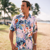 A man wearing a short-sleeve button-up Hawaiian shirt with a colorful floral print, standing outdoors on a beach with palm trees in the background.