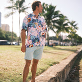 A man standing outdoors on a grassy area next to a concrete path, wearing a light pink short-sleeve button-up shirt with a colorful floral print and light blue shorts. Palm trees and a sunny outdoor setting are blurred in the background.
