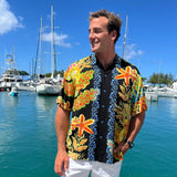 A man wearing a vibrant tropical print short-sleeve button-up shirt stands in a sunny marina with boats and blue water in the background.