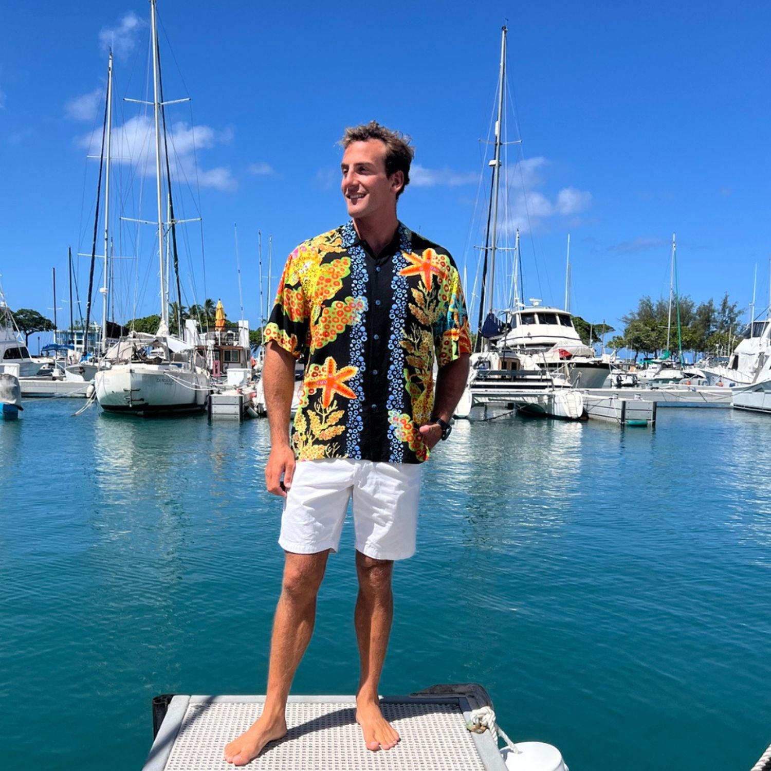 A man stands on a dock in a sunny marina wearing a colorful Hawaiian-style short-sleeve button-up shirt with yellow, orange, and blue floral patterns, paired with white shorts.
