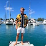 A man stands on a dock in a sunny marina wearing a colorful Hawaiian-style short-sleeve button-up shirt with yellow, orange, and blue floral patterns, paired with white shorts.