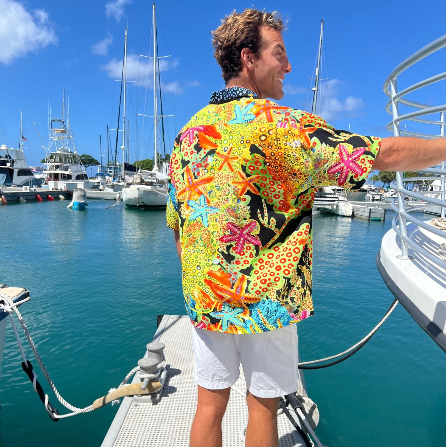 A man wearing a vibrant, multicolor short-sleeve button-up shirt with a busy tropical starfish and floral pattern, standing on a boat deck with a marina and sailboats in the background.