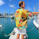 A man wearing a vibrant, multicolor short-sleeve button-up shirt with a busy tropical starfish and floral pattern, standing on a boat deck with a marina and sailboats in the background.