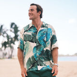 A man wearing a short-sleeve button-up shirt with a green and teal tropical leaf print on a cream background, standing on a sandy beach with palm trees and water blurred in the background.
