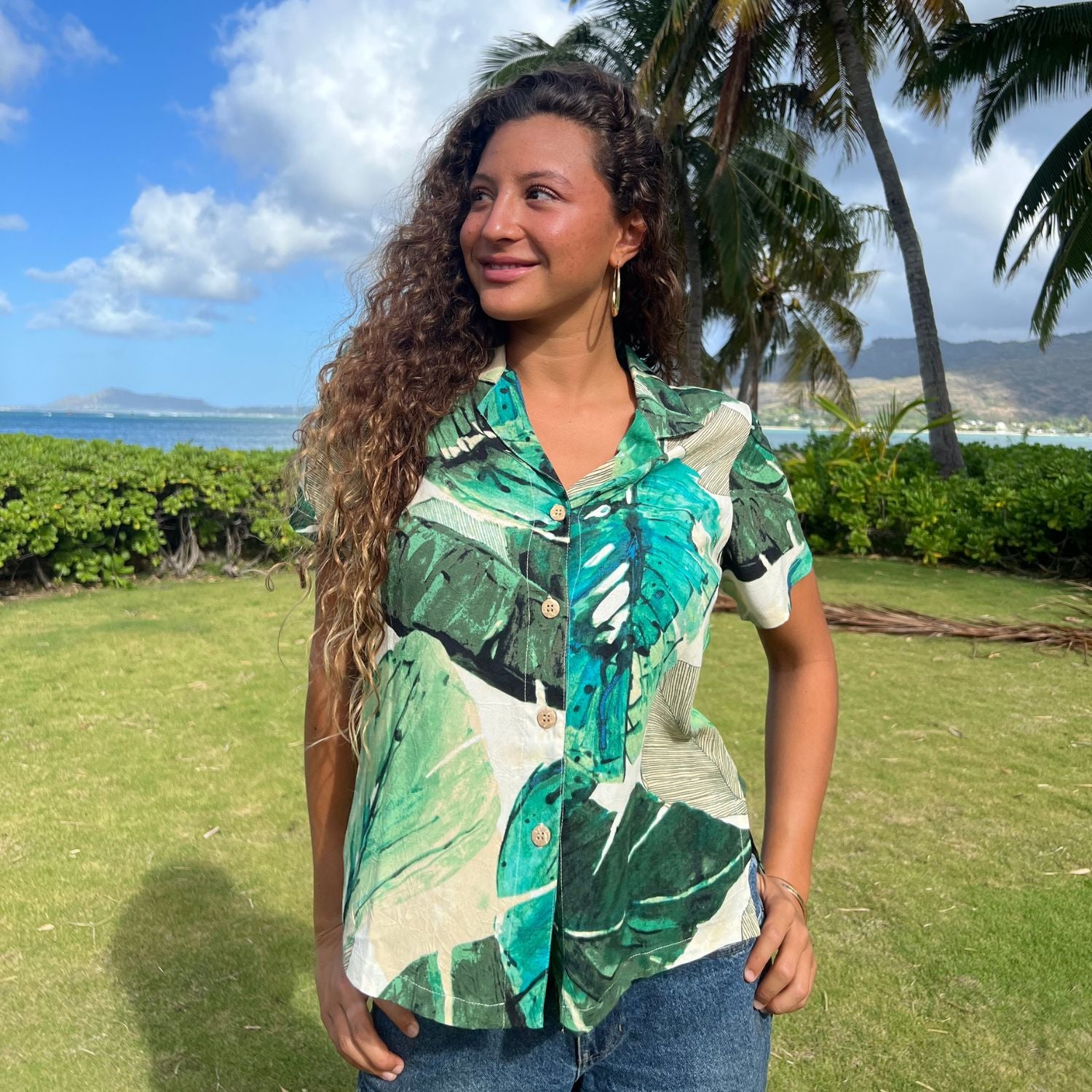 A woman wearing a short-sleeve button-up shirt with a green, teal, and white tropical leaf print, standing outdoors on a grassy lawn with palm trees and the ocean in the background.