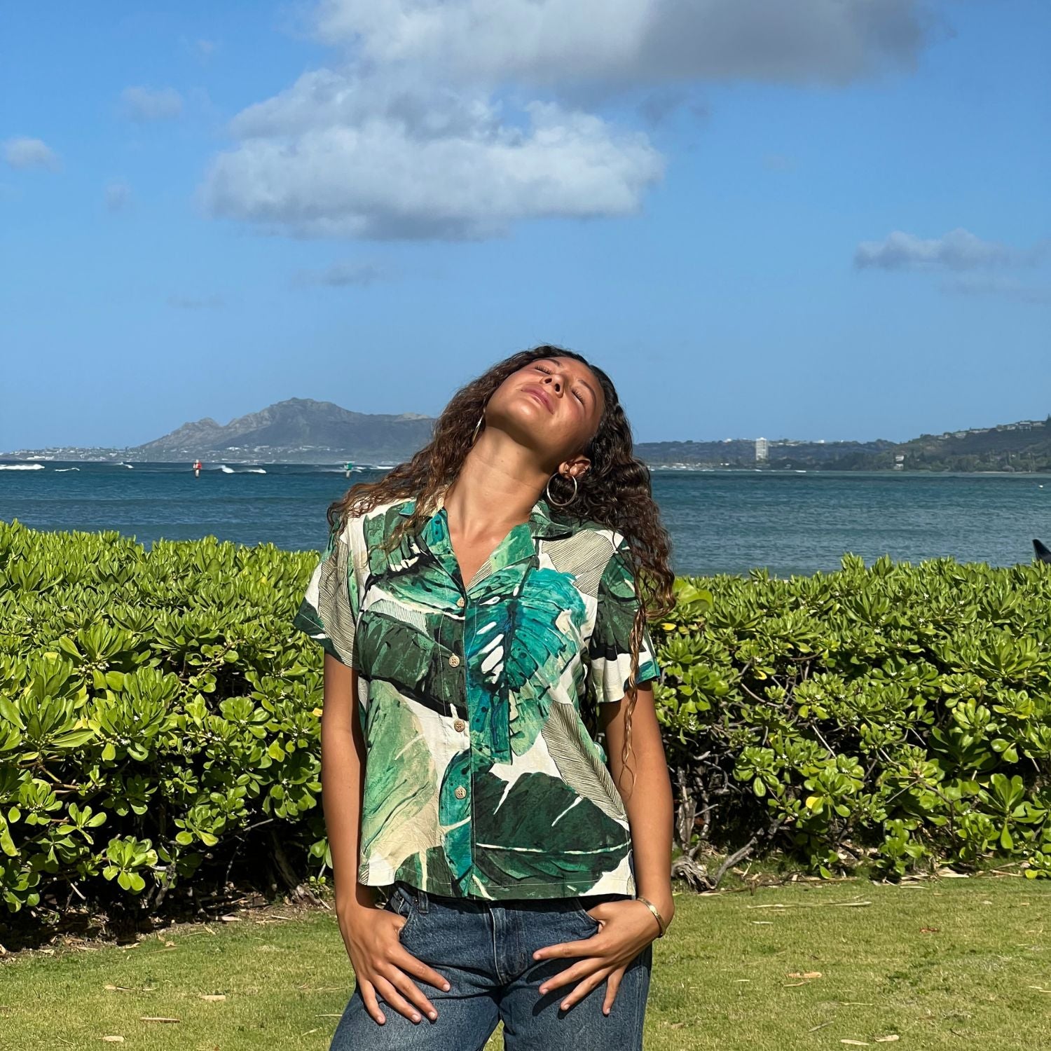A woman wearing a green and beige tropical leaf print short-sleeve button-up shirt and blue jeans stands outdoors on a grassy lawn with a green hedge, ocean, and distant mountains under a blue sky with clouds.