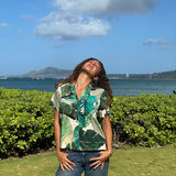 A woman wearing a green and beige tropical leaf print short-sleeve button-up shirt and blue jeans stands outdoors on a grassy lawn with a green hedge, ocean, and distant mountains under a blue sky with clouds.