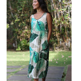 A woman wearing a sleeveless V-neck maxi dress with a green tropical leaf print on a cream background, standing outdoors in a garden setting.