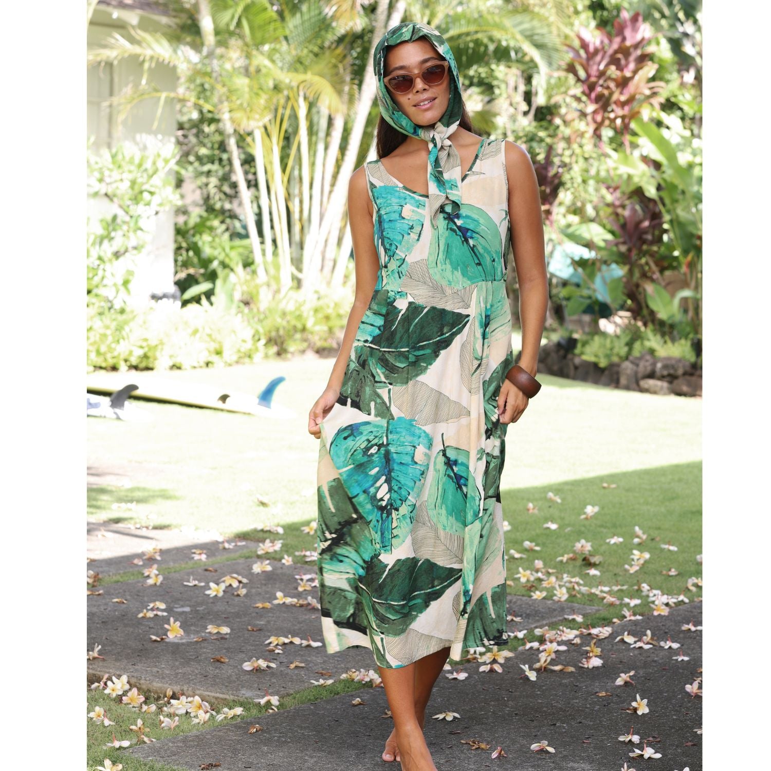 A woman wearing a tropical leaf print halter dress and matching headscarf, standing outdoors on a garden path with lush green plants in the background.