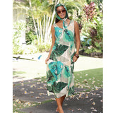 A woman wearing a tropical leaf print halter dress and matching headscarf, standing outdoors on a garden path with lush green plants in the background.