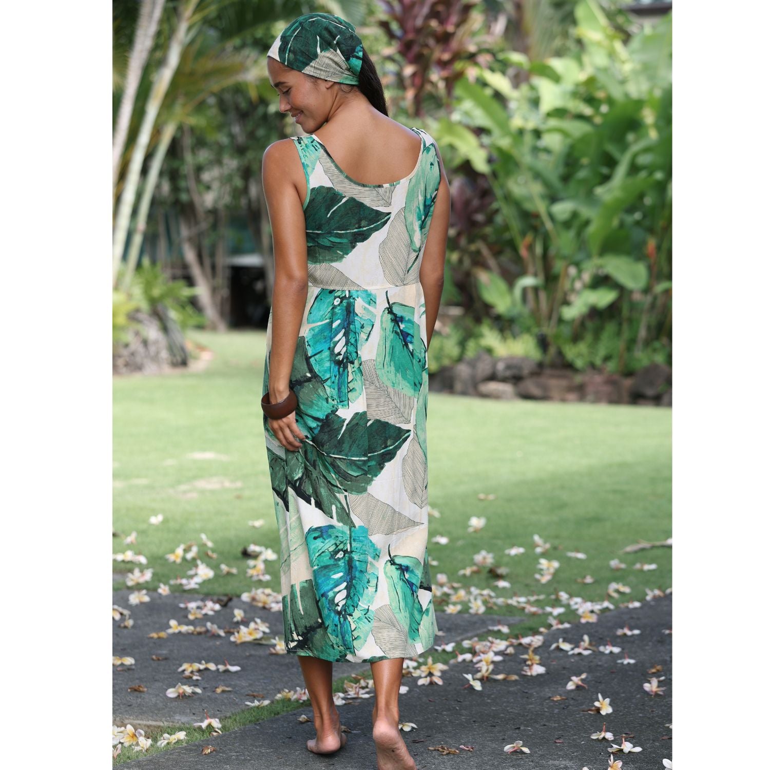 A woman wearing a sleeveless maxi dress with a green and teal tropical leaf print on a cream background, shown from the back in an outdoor garden setting.
