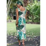A woman wearing a sleeveless maxi dress with a green and teal tropical leaf print on a cream background, shown from the back in an outdoor garden setting.