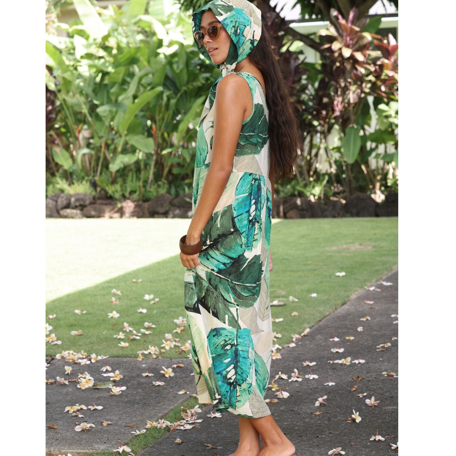 A woman wearing a sleeveless tropical leaf print maxi dress and matching headscarf, standing outdoors on a garden path.