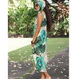 A woman wearing a sleeveless tropical leaf print maxi dress and matching headscarf, standing outdoors on a garden path.