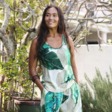 A woman wearing a sleeveless white tankini dress with a green tropical leaf print, standing outdoors in front of a white wall and green foliage.