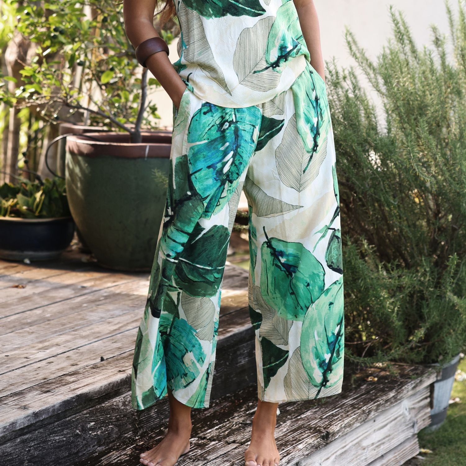 A woman wearing a matching two-piece set with a green tropical leaf print on a cream background, standing barefoot on a wooden deck with plants in the background.
