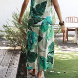 A woman wearing a sleeveless jumpsuit with a green tropical leaf print on a white background, standing barefoot on a wooden deck next to a grassy lawn.