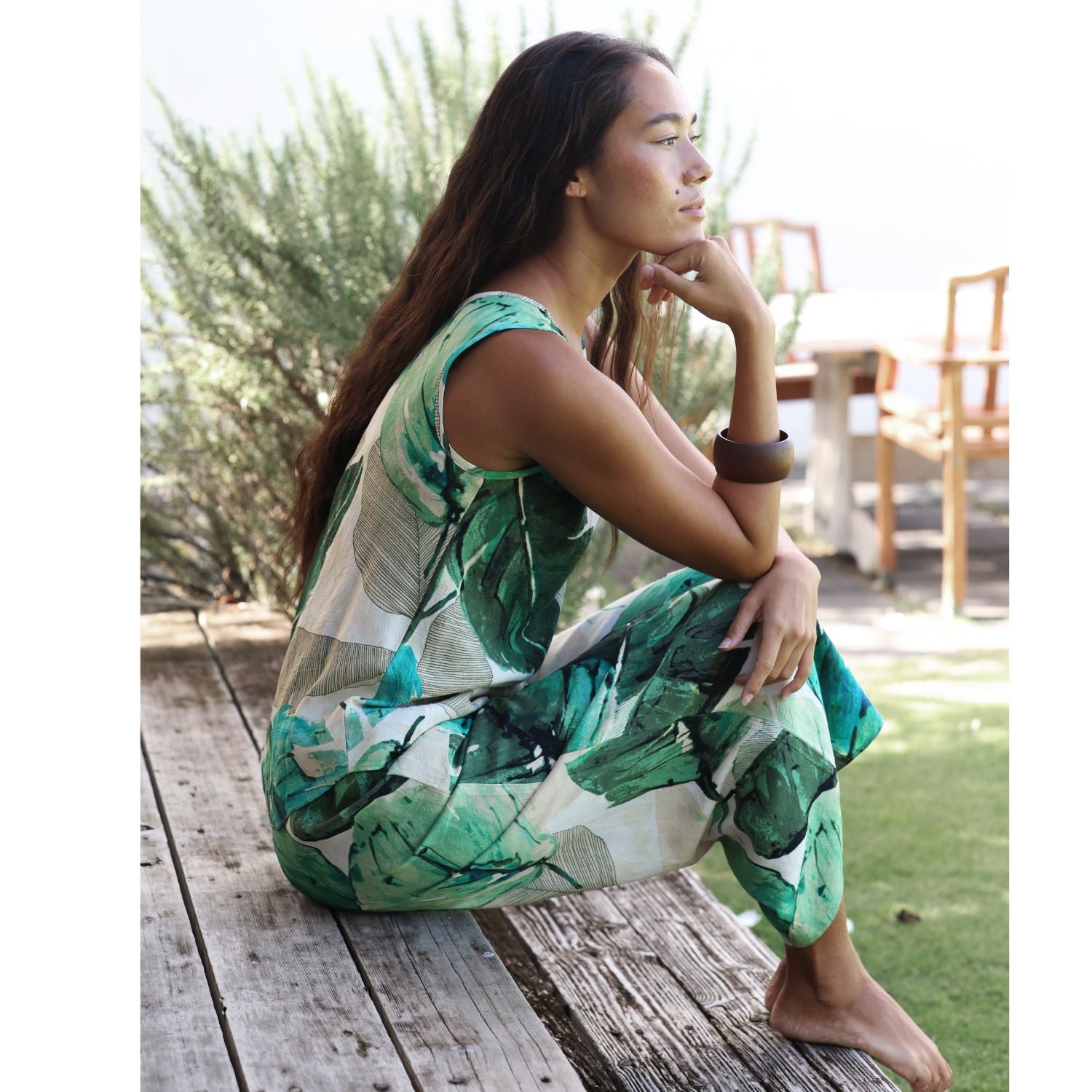 A woman sits barefoot on a weathered wooden deck outdoors, wearing a sleeveless green and white tropical leaf print dress. The background features green foliage, a wooden table, and chairs.