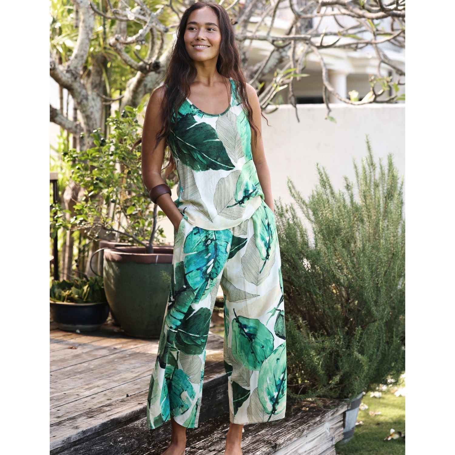 A woman modeling a sleeveless jumpsuit with a green and white tropical leaf print, standing on a wooden deck in an outdoor garden setting.