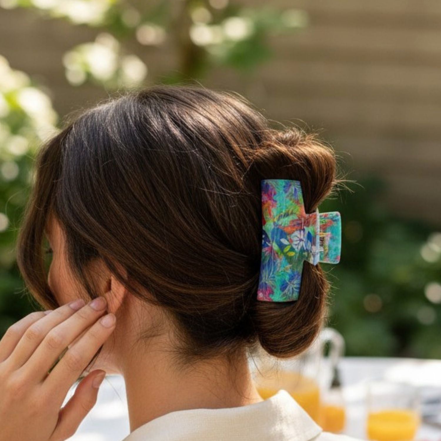A woman shown from the back wearing a colorful floral hair claw clip holding her hair in a low bun, with an outdoor blurred background.
