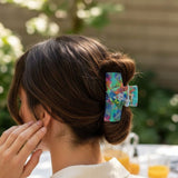 A woman shown from the back wearing a colorful floral hair claw clip holding her hair in a low bun, with an outdoor blurred background.