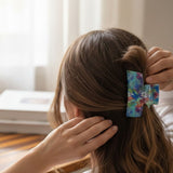 A person with long brown hair seen from the back, securing a half-up ponytail with a colorful floral hair clip. The clip has a blue, green, and purple floral pattern on a light background.