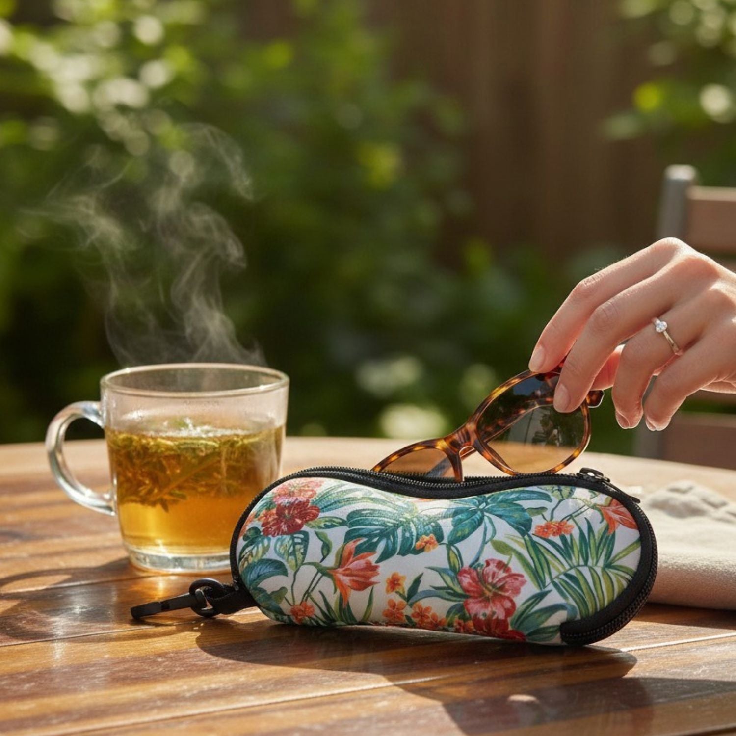 A floral-patterned sunglasses case on a wooden table, with a hand placing tortoiseshell sunglasses inside. A glass mug of tea is visible in the background, set against a blurred outdoor garden environment.