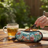 A floral-patterned sunglasses case on a wooden table, with a hand placing tortoiseshell sunglasses inside. A glass mug of tea is visible in the background, set against a blurred outdoor garden environment.
