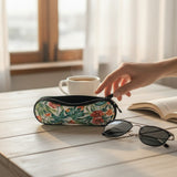 A floral-patterned zippered eyeglasses case sits on a light wooden table next to sunglasses, with a hand reaching toward it, a white coffee cup, and an open book in the background.