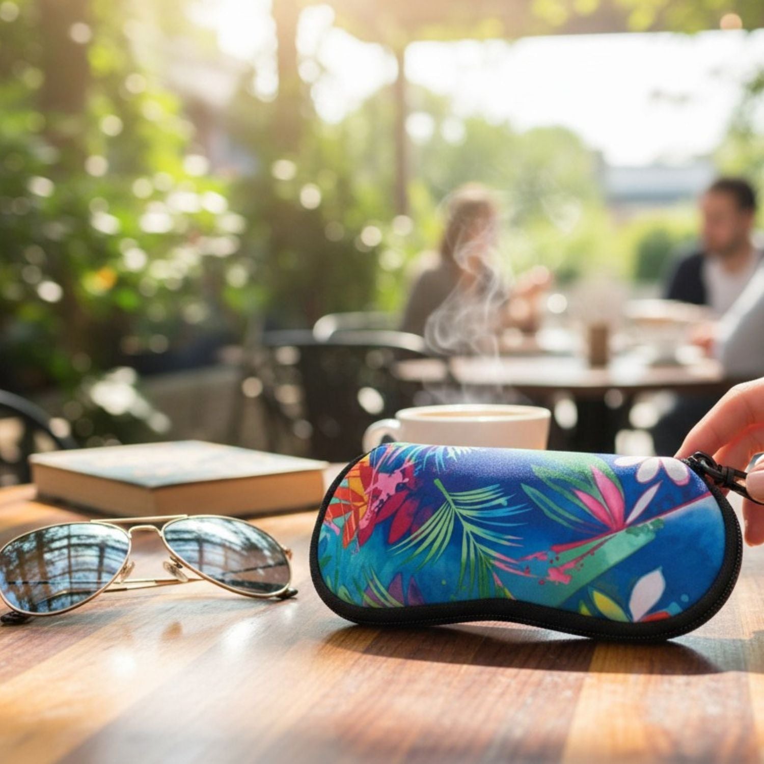 A close-up of a colorful, tropical-patterned hard sunglasses case on a wooden table, with aviator sunglasses, a book, and a coffee cup in a blurred cafe background. A hand is holding the case.