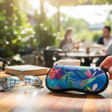 A close-up of a colorful, tropical-patterned hard sunglasses case on a wooden table, with aviator sunglasses, a book, and a coffee cup in a blurred cafe background. A hand is holding the case.