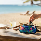 A colorful tropical-print travel pouch with a black zipper is shown on a wooden beach table, with a hand inserting sunglasses into the pouch. A straw hat, a glass of iced drink, and a blurred beach background are visible.