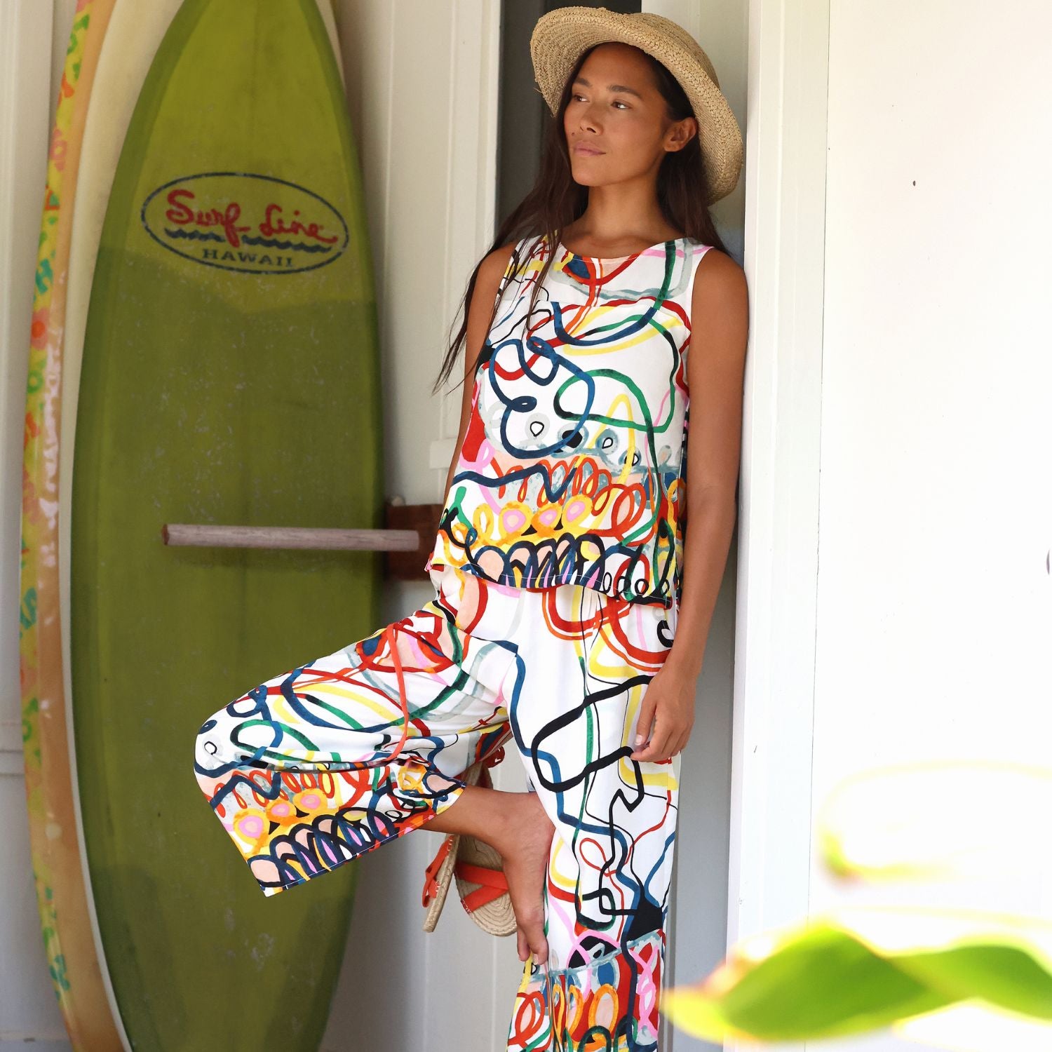 A woman wearing a sleeveless white jumpsuit with a vibrant multicolor graffiti-style abstract pattern and a straw hat, posing in front of a white door with a green surfboard in the background.