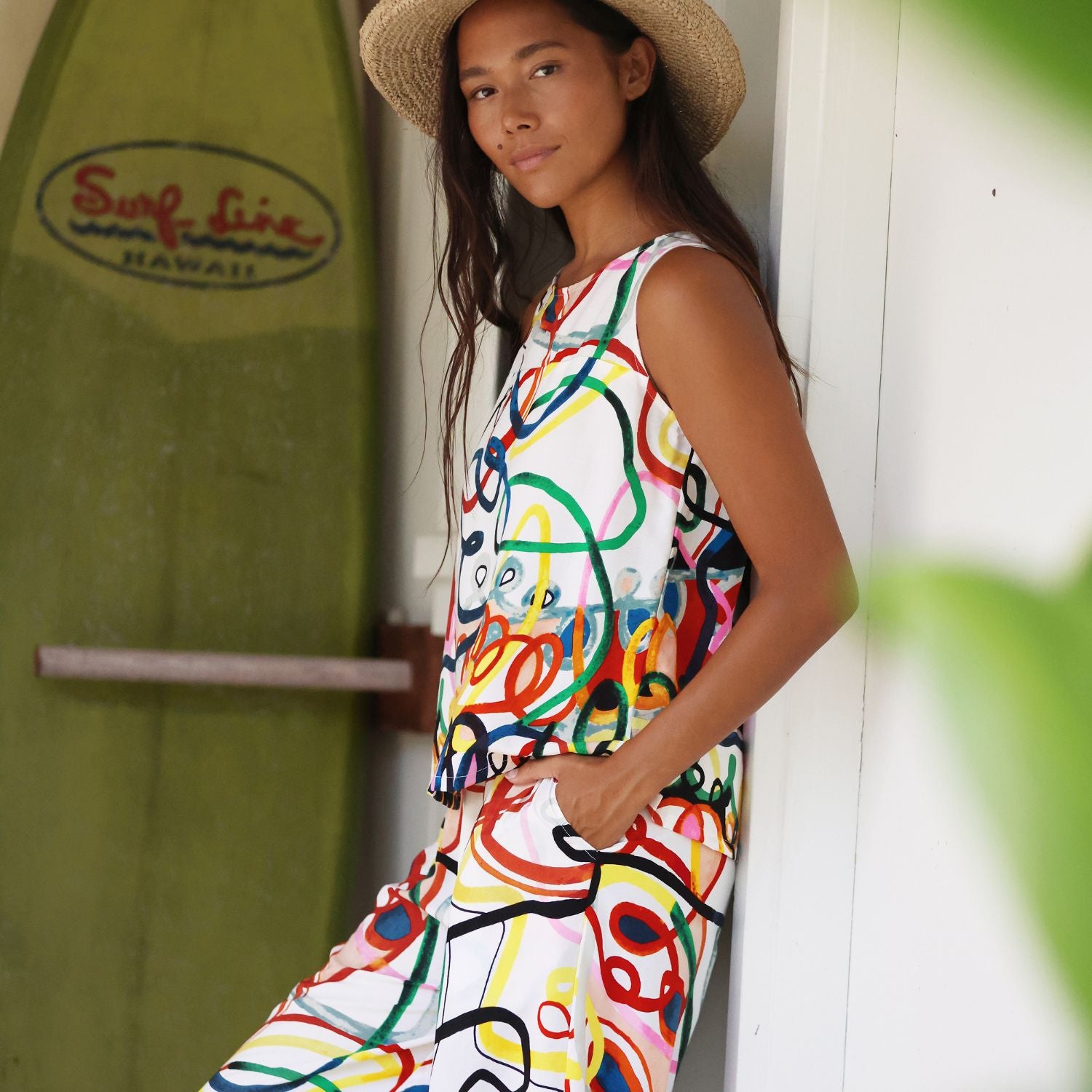 A woman wearing a sleeveless white jumpsuit with a colorful abstract line pattern leans against a white door, with a green surfboard and a blurred green leaf in the background.